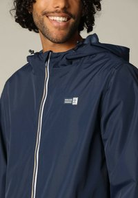 Veste à capuche bleu marine avec une texture lisse, dotée d'une fermeture éclair blanche et d'un petit logo brodé sur la poitrine. Design léger et décontracté.