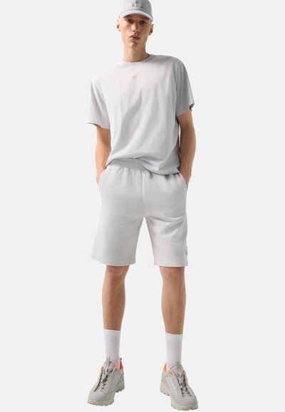 Jeune homme en t-shirt gris clair, short assorti, chaussettes blanches, baskets grises avec des accents orange et une casquette gris clair, debout les mains dans les poches.