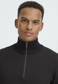 Čierny pletený sveter s vysokým golierom a zlatým zipsom. Obsahuje textúrovaný vzor, priliehavý strih a čistý, moderný vzhľad.