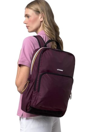 Donna bionda con camicia rosa e pantaloni bianchi che indossa un grande zaino marrone scuro con tasca frontale con cerniera e dettagli a righe sulle spalline.