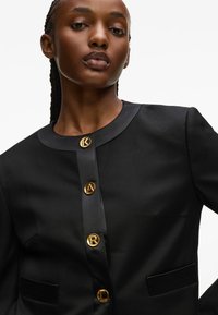 Fekete blézer kerek nyakkivágással, arany logós gombokkal és oldalsó zsebekkel. Sima anyag, testreszabott kialakítással és tiszta vonalakkal.