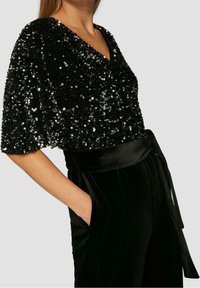 Svart paljettopp med V-ringning och fladdermusärmar; matchad med högmidjade svarta sametbyxor med knytmidja och sidfickor.