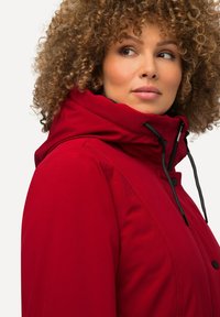 Red waterproof jacket with a high collar, adjustable hood, black drawstrings, and snap buttons. Smooth texture and fitted design.