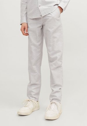 Personne portant un pantalon gris clair et des baskets blanches, debout avec une main dans la poche, sur un fond uni.