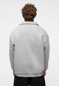 Grijze oversized sweatshirt met een opgerold kraag, verlaagde schouders en geribbelde mouwen, gemaakt van zachte stof, gedragen over donkere broeken.