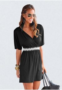 Woman wearing black dress with white patterned waistband, aviator sunglasses, large feather earrings, gold bracelets, holding black woven bag.