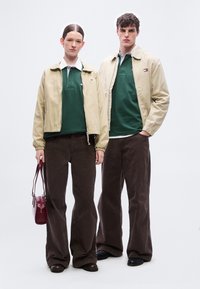 Δύο μοντέλα στέκονται δίπλα δίπλα φορώντας μπεζ σακάκια πάνω από πράσινα πόλο πουκάμισα, καφέ παντελόνες με φαρδιά πόδια· το ένα κρατά μια κόκκινη τσάντα χειρός.