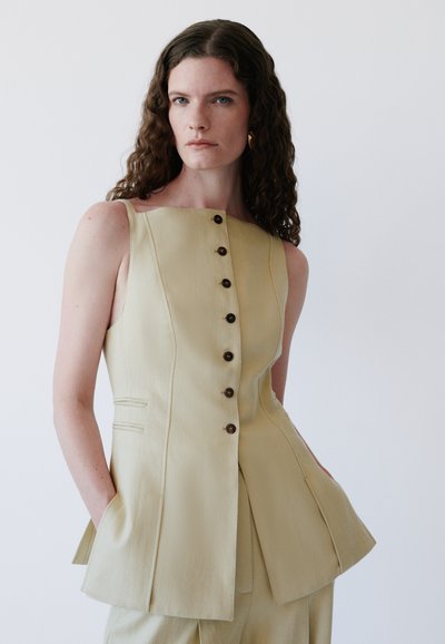 Γυναίκα με μακριά σγουρά μαλλιά φοράει μπεζ αμάνικο τοπ με κουμπιά και πλαϊνές τσέπες, στέκεται μπροστά σε απλό ανοιχτό φόντο.