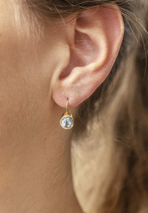Orecchino a cerchio in oro con una pietra preziosa circolare di colore azzurro chiaro. La pietra è sfaccettata, riflettendo la luce, ed è attaccata a un sottile filo d'oro.