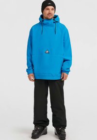 Uomo che indossa una giacca con cappuccio di un blu vivace, pantaloni neri, scarponi da sci neri e un berretto nero, in piedi contro uno sfondo chiaro e uniforme.