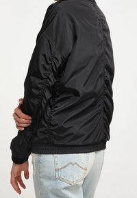 Personne portant une veste noire en nylon froncée et un jean délavé bleu clair, tenant un bras croisé sur le corps.