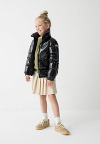 Giacca a vento nera lucida con colletto alto e polsini elastici, abbinata a un maglione verde chiaro, una gonna beige plissettata e sneaker color miele.