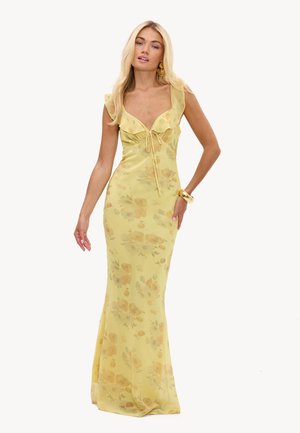 Femme portant une longue robe jaune ajustée à motifs floraux avec des épaules volantées, accessoirisée de boucles d'oreilles en or et d'un grand bracelet en or.