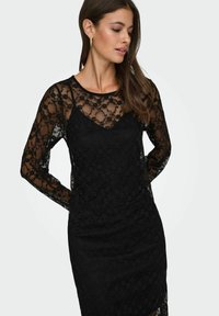 Czarna koronkowa sukienka o dopasowanym kroju, z długimi przezroczystymi rękawami i kwiatowymi wzorami. Dekolt w serek z gładką wewnętrzną warstwą.
