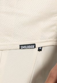 Top deportivo color crema con una tela texturizada y transpirable, que presenta una etiqueta negra de SMILODOX cosida en el dobladillo.