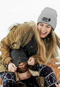 Gray knit beanie with a textured ribbed design and a printed owl graphic. Snowflakes are visible in a winter outdoor setting.