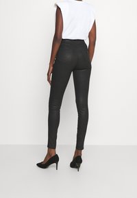 Mujer vestida con jeans ajustados de cintura alta negros, blusa sin mangas blanca y zapatos de tacón negros, de pie contra un fondo liso.