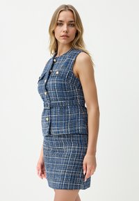 Abito in tweed blu, strutturato con una silhouette attillata, design senza maniche e bottoni dorati. La gonna è sagomata a matita.