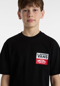 Camiseta de manga corta negra de algodón que presenta un logo de Vans en blanco y rojo en el lado izquierdo del pecho. Textura suave, cuello redondo.