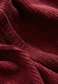 Tissu en velours côtelé bordeaux profond avec des rainures verticales douces et surélevées, ainsi que des plis légers créant des ombres et des reflets texturés et en couches.