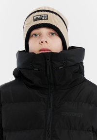 Zwarte gewatteerde jas met hoge kraag, ritssluiting aan de voorkant en trekkoorden. Beige geribbelde beanie met zwarte strepen en een logopatch.