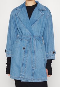 Džínový trenčkot světle modré barvy se širokými klopami, páskem v pase a zapínáním na knoflíky na manžetách. Má volný střih a výrazné šicí detaily.