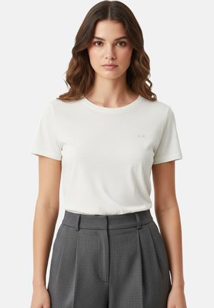 Giovane donna con capelli castani ondulati che indossa una maglietta bianca a maniche corte infilata in pantaloni grigi a pieghe a vita alta.