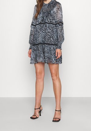 Schwarz-weißes Zebra-Muster-Kleid mit einem gerafften Ausschnitt, transparentem Stoff, langen Ärmeln und einem gestuften Rock. Kombiniert mit schwarzen Absatzsandalen.