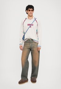 Pull en tricot crème avec "BONSAI" et le numéro 10 en rouge, accentué de rayures teal. Porté avec un jean bleu décoloré et usé ainsi que des chaussures marron.