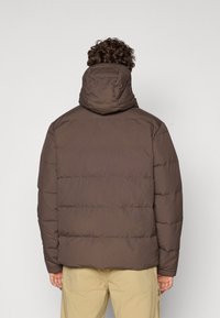 Brun pufferjakke med hætte, der har horisontale syninger og en blød, quilted tekstur. Designet er enkelt og strømlinet.