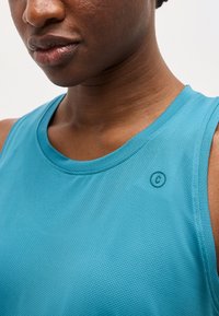 Κοντινό πλάνο ενός ατόμου που φοράει μπλε αμάνικο τοπ με υφή και μικρό κυκλικό λογότυπο κοντά στον ώμο, σε ανοιχτό φόντο.