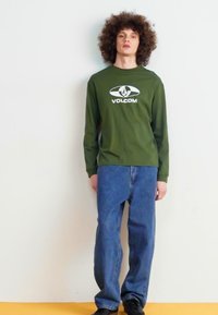 Grønn langermet skjorte med en hvit "VOLCOM" logo, kombinert med blå vide jeans og svarte sko, mot en nøytral bakgrunn.