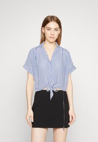 Chemise à rayures bleues et blanches à manches courtes avec un devant noué, associée à une mini-jupe noire avec une fermeture éclair sur le côté.