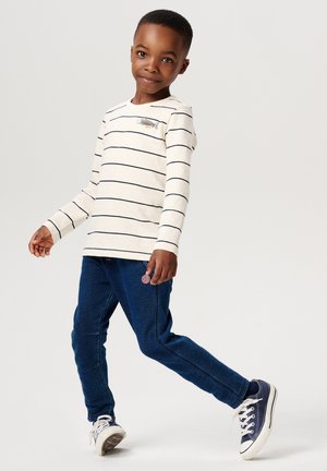 Weißes Langarmhemd mit marineblauen Streifen, versehen mit einer Fischbordüre, kombiniert mit blauen Jeans. Sneakers runden das Outfit ab.