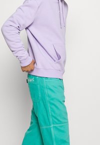 Vaaleanvioletti huppari, jossa etutasku ja kiristysnauhat, yhdistetty sinivihreisiin housuihin, joissa on kontrastiväriset saumat ja sivussa merkintä.