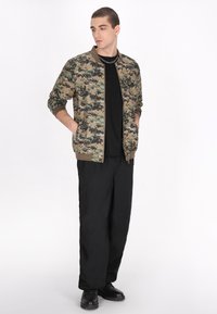 Camouflage bomberjakke i khaki, grønne og sorte nuancer, med ribbede ærmer og kant. Sort t-shirt, løse sorte bukser og sorte sko.