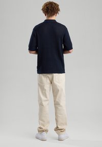 Navy kauluspaita, jossa lyhyet hihat ja teksturoitu kuvio, yhdistettynä beigeihin housuihin ja valkoisiin tenniskenkiin. Näkyy takanäkymä.