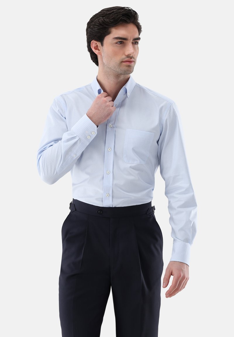 Uomo che sistema il colletto di una camicia azzurro chiaro con bottoni, indossa pantaloni scuri plissettati, guarda di lato contro uno sfondo semplice.