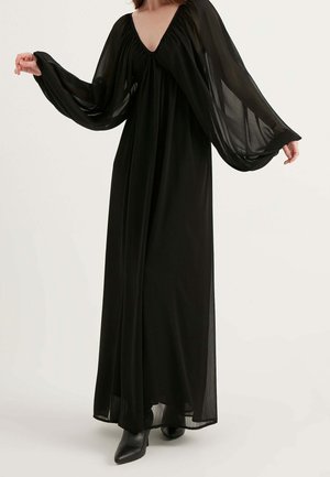 Kobieta w długiej czarnej sukni z prześwitującymi, obszernymi rękawami typu biskup oraz czarnych, szpiczastych botkach na białym tle.