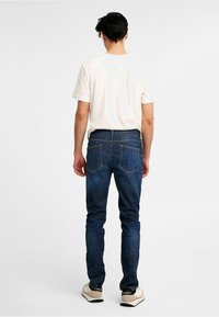 Blå denimjeans med slim fit, femfickorsdesign, synliga stygn och en enkel vit t-shirt sedd bakifrån.