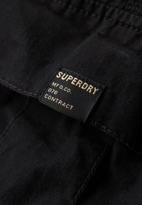 Étiquette de vêtement en tissu noir avec texte doré indiquant "SUPERDRY MFG.CO. 076 CONTRACT" cousue sur un vêtement sombre.