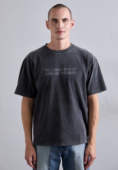 Uomo che indossa una maglietta nera sbiadita con il testo "Art Department 1995-08 MISBHV", abbinata a un paio di jeans bicolore blu chiaro e scuro.