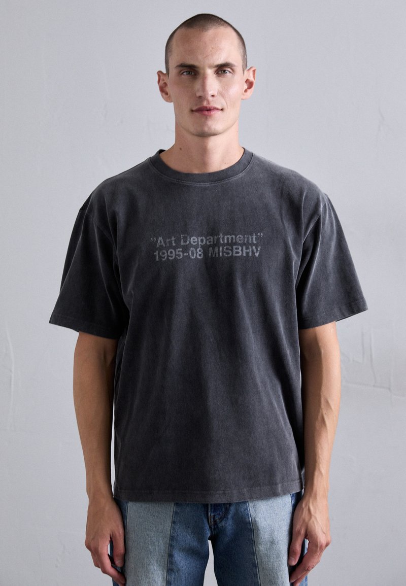 Mann trägt ein verblasstes schwarzes T-Shirt mit dem Text "Art Department 1995-08 MISBHV", kombiniert mit zweifarbigen hell- und dunkelblauen Jeans.