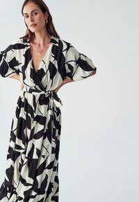 Haljina s uzorkom u crno-bijeloj kombinaciji, s širokim rukavima, dubokim V izrezom i vezanim strukom. Izrađena od lagane tkanine, s višeslojnim suknjom.