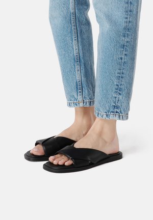 Pėdos, avinčios šviesiai mėlynas džinsines kelnes ir juodus odinius šlepetes su kryžminėmis juostelėmis, ant balto fono.