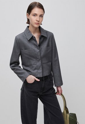 Młoda kobieta z krótkimi włosami, ubrana w szarą krótką kurtkę i czarne spodnie, trzyma zieloną torebkę na jednolitym tle.