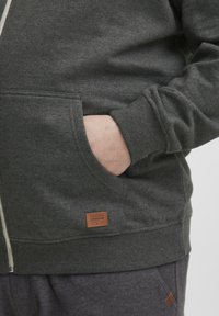 Tamno siva dukserica na zip, napravljena od mekanog materijala, sa oznakom na prednjoj strani i prednjim džepom, opuštenog kroja.