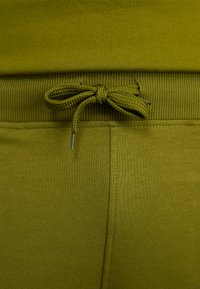 Leggings verde oliva con un elastico testurizzato in vita dotato di cordini. Il tessuto è liscio con una leggera lucentezza e cuciture strutturate.