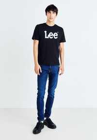 Melna īsām piedurknēm t-kreklu ar baltu "Lee" logo, kombinēts ar ziliem šauriem džinsiem un melniem sporta apaviem, stāvot pret baltu fona.