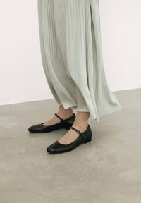 Zapatos planos de cuero negro con puntera redondeada y tira en el tobillo. El diseño presenta un tacón bajo y una textura suave.